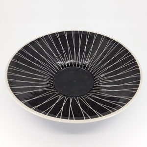 Può includere: Una ciotola in ceramica nera con un design geometrico bianco. La ciotola ha un bordo largo e un centro leggermente rialzato.