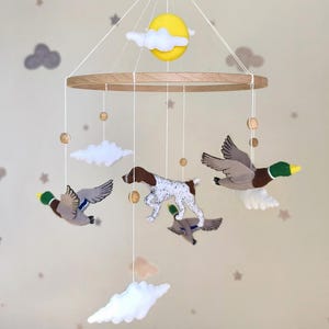 May include: A hanging mobile featuring a circular wooden frame suspended by white strings. Attached are felt figures of a brown and white spotted dog, three mallard ducks in flight, white clouds, and a yellow sun, accented with small wooden beads.