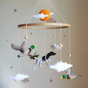 May include: A hanging nursery mobile featuring a circular wooden frame with felt figures suspended by white strings. The mobile includes a bright orange sun, white clouds, two mallard ducks in flight, and a black and white spotted hunting dog.