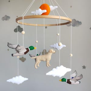 Pode incluir: Móvel de berço com um aro de madeira, patos de feltro, um cão, nuvens e um sol laranja. Os patos têm detalhes verdes e castanhos, o cão é bege e as nuvens são brancas. Suspenso por fios brancos.