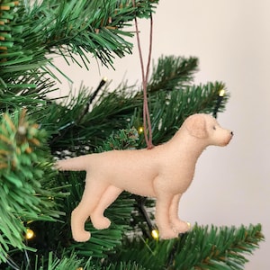 May include: A light tan Labrador Retriever dog ornament hanging on a green Christmas tree. The dog is made of felt and has a brown string for hanging. The tree has small yellow lights.