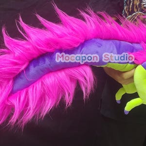 Op de afbeelding: Een pluche draken speelgoed met een paars lichaam, groene voeten en een lange, roze, pluche staart. De draak wordt vastgehouden door een hand.