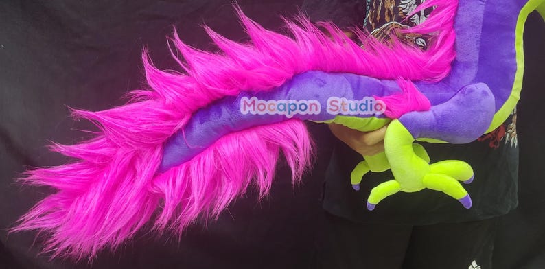Op de afbeelding: Een pluche draken speelgoed met een paars lichaam, groene voeten en een lange, roze, pluche staart. De draak wordt vastgehouden door een hand.