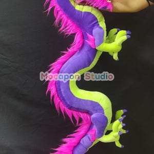 Op de afbeelding: Een groot pluche draken speelgoed met een paarse en groene body, roze vacht en gele accenten. De draak heeft een wijd open mond en grote, witte ogen.
