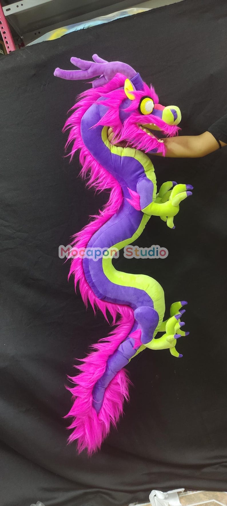 Op de afbeelding: Een groot pluche draken speelgoed met een paarse en groene body, roze vacht en gele accenten. De draak heeft een wijd open mond en grote, witte ogen.