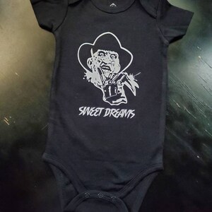 May include: Black baby bodysuit with a silver graphic of a horror movie character wearing a hat and glove with blades. The text "SWEET DREAMS" is printed above the character's image. Short-sleeved, one-piece garment.
