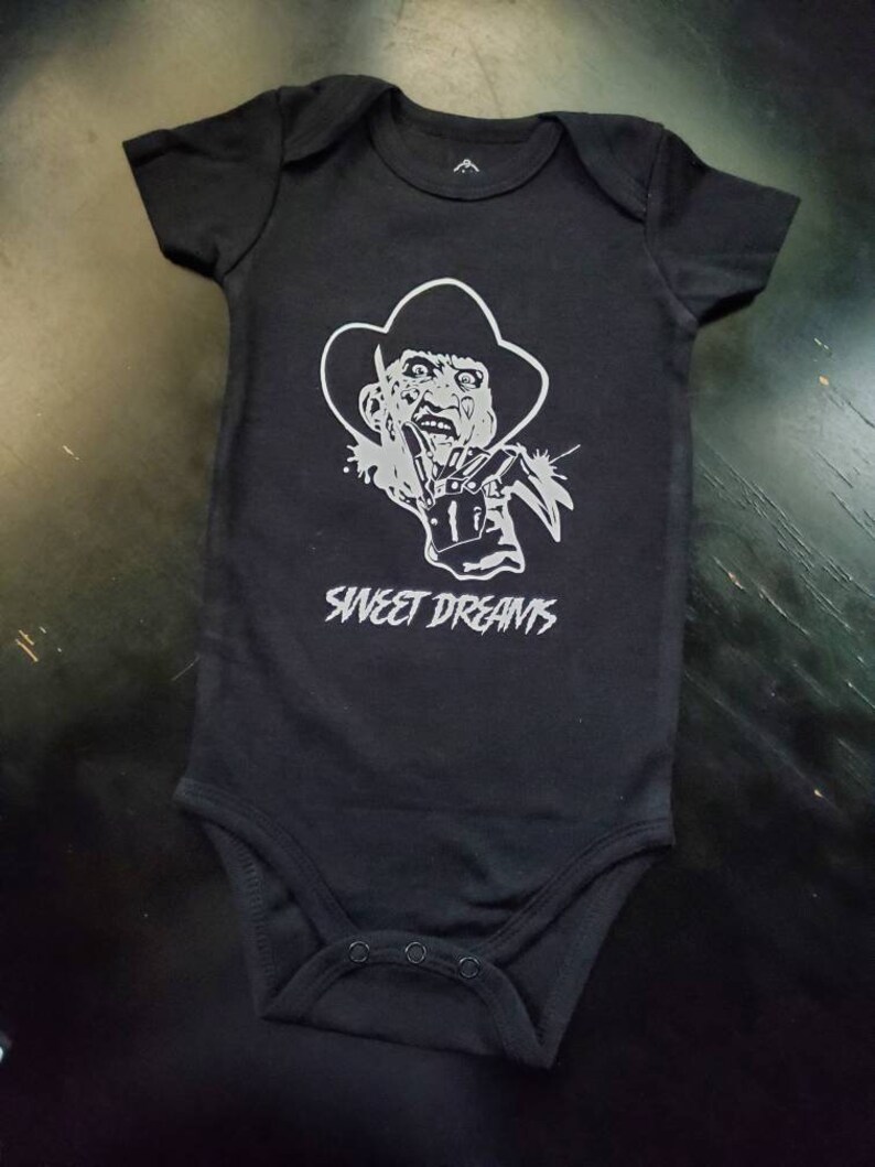 May include: Black baby bodysuit with a silver graphic of a horror movie character wearing a hat and glove with blades. The text "SWEET DREAMS" is printed above the character's image. Short-sleeved, one-piece garment.