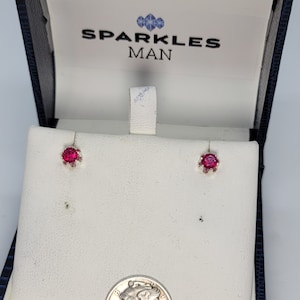 May include: A pair of red gemstone earrings in a blue velvet jewelry box with a gold clasp. The box has the words "Sparkles Man" printed on the inside.