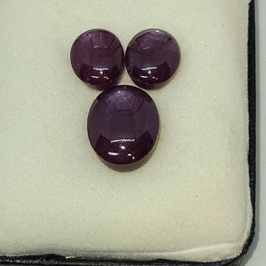 May include: Three oval-shaped gemstones in a velvet-lined box. The gemstones are a deep purple color and have a glossy finish.