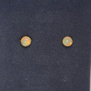 May include: A pair of opal stud earrings. Each earring features a round, iridescent opal stone set in a gold-colored bezel. The opals display a mix of colors, including white, orange, and green, against a dark blue background.