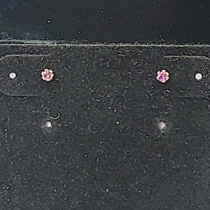 May include: A pair of small stud earrings. Each earring features a small, round, pink gemstone set in a gold-colored metal. The earrings are displayed on a dark, textured surface.