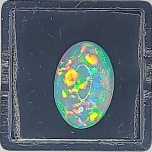 May include: An oval-shaped opal gemstone with a vibrant display of colors, including green, yellow, and orange. The opal is set against a dark background, showcasing its iridescent qualities. The gemstone is a natural, untreated opal.
