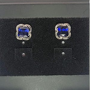 May include: A pair of square-cut, dark blue gemstone stud earrings. Each earring features a silver-toned, openwork frame. The earrings are displayed on a black velvet surface.