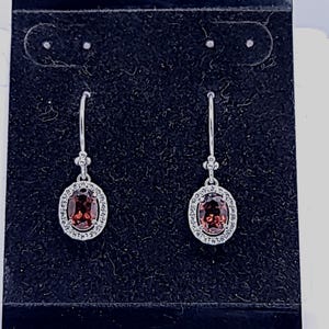 May include: A pair of silver dangle earrings with oval-shaped red gemstones surrounded by a halo of smaller clear gemstones.