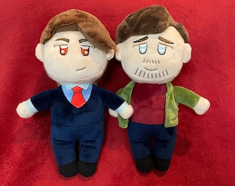 May include: Two plush dolls of men, each with brown hair and embroidered facial features. One doll wears a navy suit with a red tie, the other a green jacket over a burgundy shirt. Both dolls have black shoes and are on a red background.
