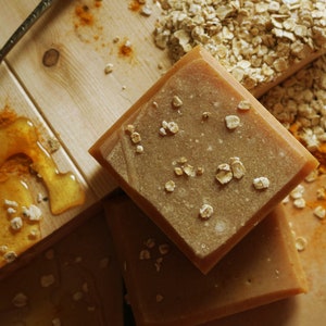 May include: Three bars of handmade soap with a light brown color. The soap is topped with oats and has a honey-like texture. The soap is sitting on a wooden surface with honey and turmeric.