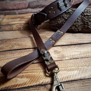 May include: A brown leather dog collar and lead set. The collar features a metal Thor's hammer emblem and brass rivets. The lead has a matching emblem and a brass clasp. The set is displayed on a wooden surface.