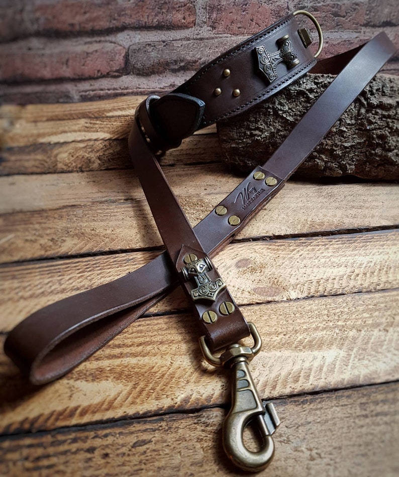 May include: A brown leather dog collar and lead set. The collar features a metal Thor's hammer emblem and brass rivets. The lead has a matching emblem and a brass clasp. The set is displayed on a wooden surface.