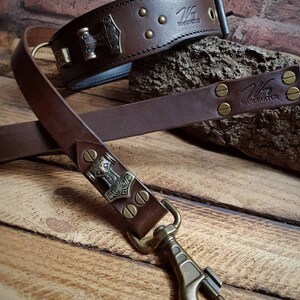 May include: A brown leather dog collar and lead set. The collar features a metal Thor's hammer emblem and brass studs. The lead has a matching design with a brass clasp. The brand name "Wiking Manufaktur" is visible on both items.