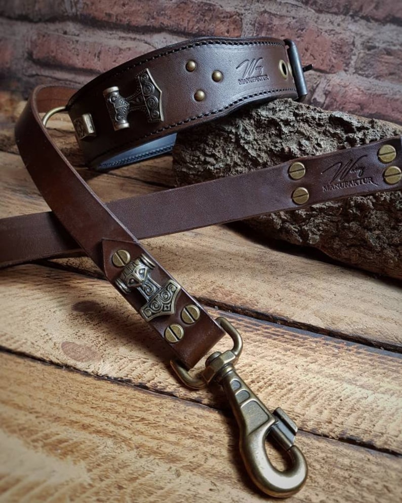 May include: A brown leather dog collar and lead set. The collar features a metal Thor's hammer emblem and brass studs. The lead has a matching design with a brass clasp. The brand name "Wiking Manufaktur" is visible on both items.