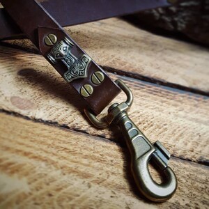 May include: A close-up of a brown leather strap with a metal clasp and a decorative plate featuring a hammer design. The strap is secured with brass-coloured rivets and hardware, suggesting a handcrafted item.