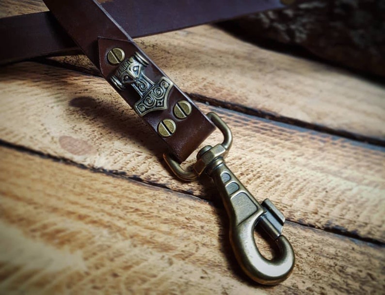 May include: A close-up of a brown leather strap with a metal clasp and a decorative plate featuring a hammer design. The strap is secured with brass-coloured rivets and hardware, suggesting a handcrafted item.