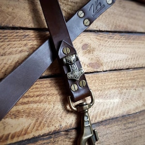 May include: A brown leather strap with a bronze clasp and a decorative metal piece featuring a hammer design. The strap is riveted and stamped with the text "W MARZAKTUR". The background is a wooden surface.