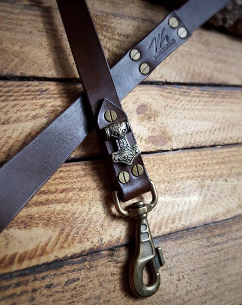 May include: A brown leather strap with a bronze clasp and a decorative metal piece featuring a hammer design. The strap is riveted and stamped with the text "W MARZAKTUR". The background is a wooden surface.