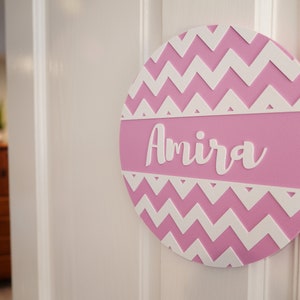 May include: Pink and white chevron patterned round wooden sign with the name Amira written in white.
