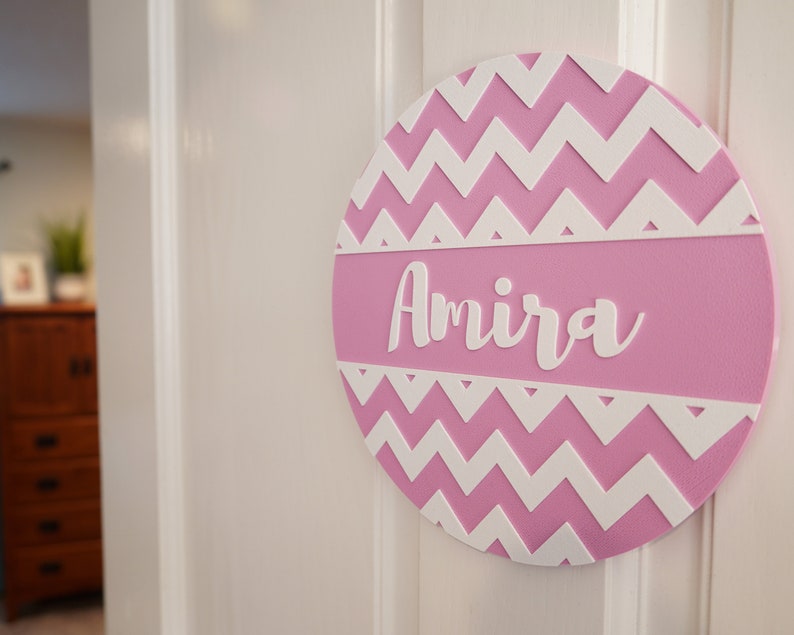 May include: Pink and white chevron patterned round wooden sign with the name Amira written in white.