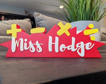 Könnte beinhalten: Ein rotes und gelbes Schreibtischschild mit dem Text "Miss Hodge" und mathematischen Symbolen: Plus, Minus, Multiplizieren und Dividieren.