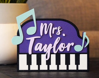 May include: Purple piano-shaped sign with the text "Mrs. Taylor" in white and light purple lettering. Two light blue musical notes are on either side of the sign.