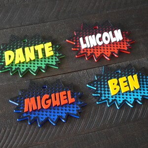May include: Four colorful, personalized name tags with a comic book style design. The names are Dante, Lincoln, Ben, and Miguel. The tags are made of a thick, durable material and have a glossy finish.