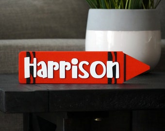 Puede incluir: Un letrero de madera rojo en forma de crayón con el nombre "Harrison" en letras blancas. El letrero tiene rayas negras en el lateral.