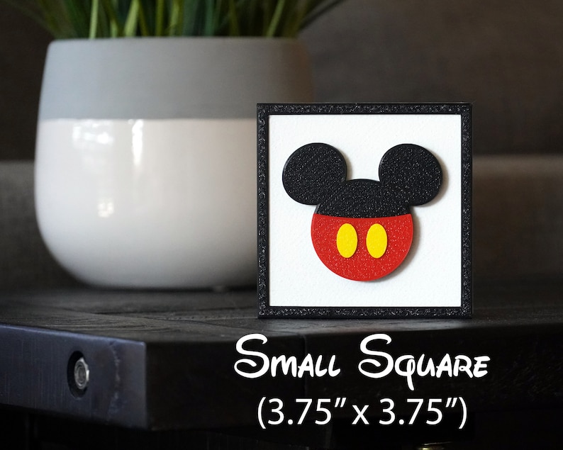 May include: A black framed square wall art piece with a white background featuring a red and black Mickey Mouse head with yellow circles for eyes. The text "Small Square (3.75" x 3.75")" is printed below the image.