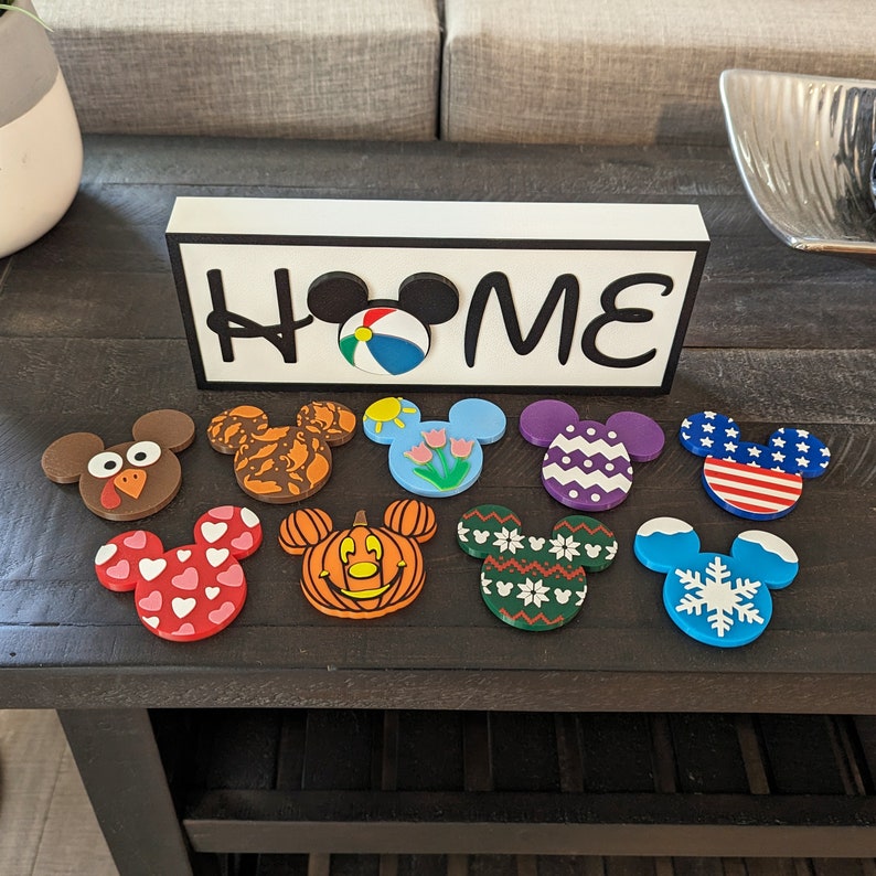 May include: A white wooden sign with the word "HOME" in black letters and a Mickey Mouse head with a beach ball in the middle. There are 10 colorful Mickey Mouse head magnets with different designs, including a turkey, a pumpkin, a snowflake, and a heart.