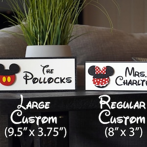 May include: Two custom wooden signs with Mickey Mouse and Minnie Mouse characters. The larger sign is 9.5 inches by 3.75 inches and reads "The Pollocks". The smaller sign is 8 inches by 3 inches and reads "Mrs. Charlton".