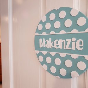 May include: A light blue round door sign with white polka dots and the name "Kenzie" in white lettering.