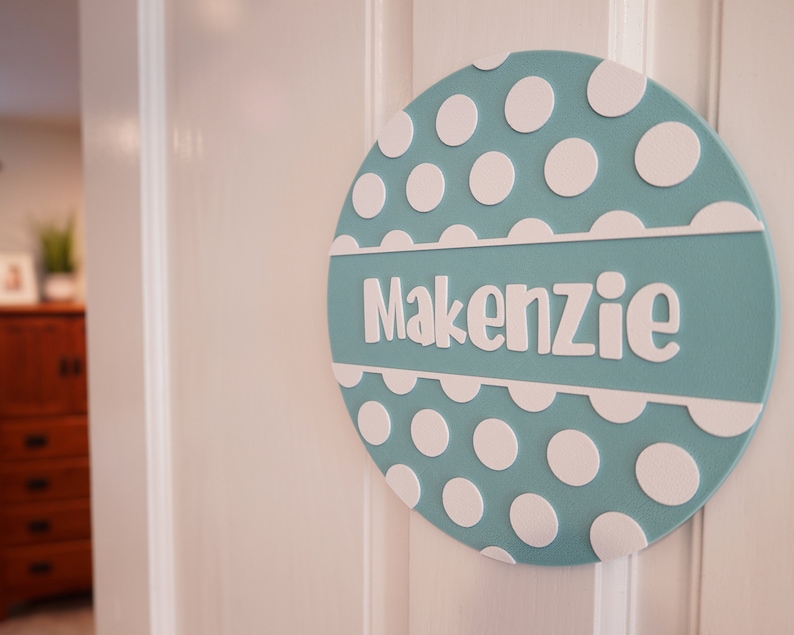 May include: A light blue round door sign with white polka dots and the name "Kenzie" in white lettering.