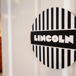 May include: A black and white striped round sign with the word "LINCOLN" in white letters on a black background.