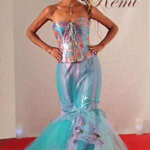 May include: A woman wearing a blue and purple mermaid-style gown with a floral print corset top. The gown has a long train and is made of tulle. The woman is standing on a red carpet. The text "Upscale Gowns" and "Remi" is visible in the background.