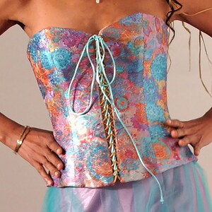 May include: A strapless corset with a colorful floral pattern and a turquoise lace-up front. The corset is made of a silky fabric and has a fitted silhouette.