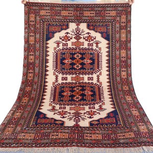 Puede incluir: Una alfombra rectangular con un campo central crema y patrones geométricos detallados en tonos rojos, azules y marrones. La alfombra presenta un borde detallado y flecos a lo largo de los bordes, mostrando artesanía tradicional.