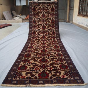 May include: A long, rectangular rug with a detailed pattern. The rug features a cream-coloured central field with repeating geometric and floral motifs in red, brown, and green. The border is a dark brown with floral designs. The rug is displayed outdoors.