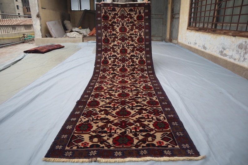 May include: A long, rectangular rug with a detailed pattern. The rug features a cream-coloured central field with repeating geometric and floral motifs in red, brown, and green. The border is a dark brown with floral designs. The rug is displayed outdoors.