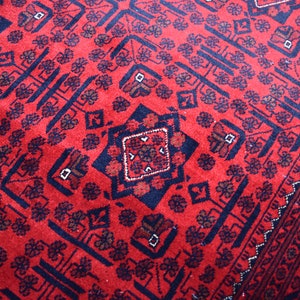 May include: Close-up of a red rug with a detailed geometric pattern. The rug features a repeating design of dark blue shapes and floral motifs. The border has a decorative pattern.