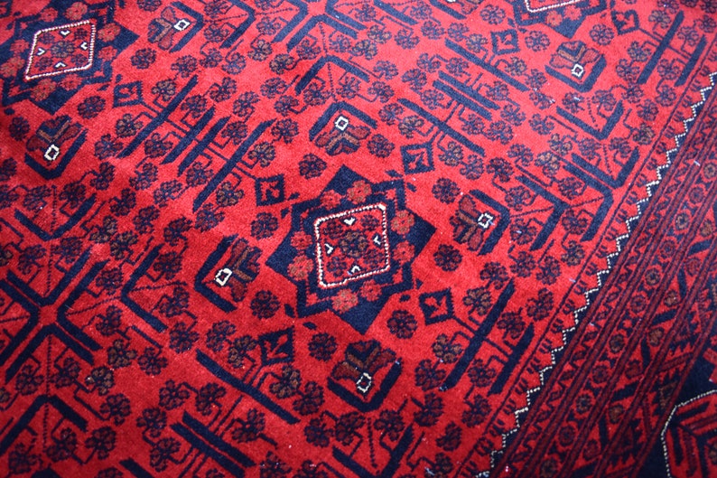 May include: Close-up of a red rug with a detailed geometric pattern. The rug features a repeating design of dark blue shapes and floral motifs. The border has a decorative pattern.