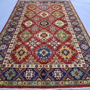 May include: A rectangular, hand-knotted rug with a central field of red, featuring floral and geometric patterns in blue, green, yellow, and cream. The rug has a detailed border in blue and green, with a cream-coloured inner border. The rug is likely made of wool.