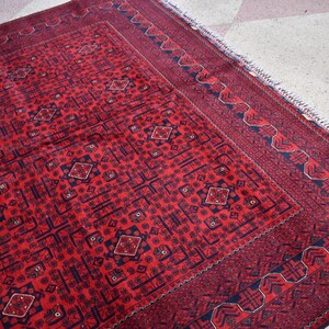 May include: A rectangular, red rug with a detailed geometric pattern. The rug features a central field of repeating diamond and square motifs in a darker shade of red and navy blue. A navy blue border with a repeating design frames the rug.