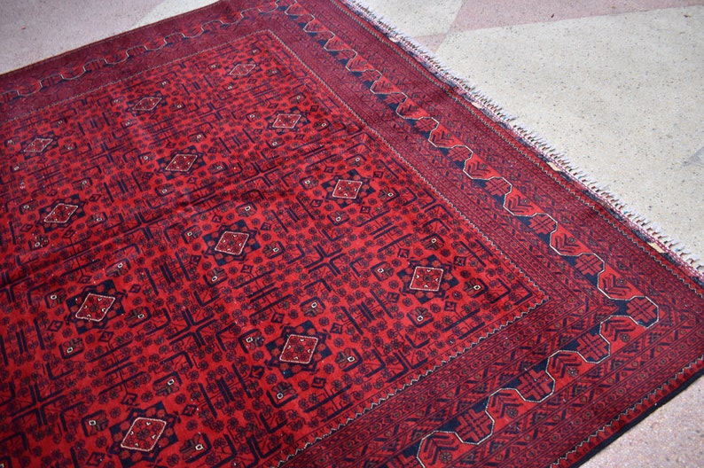May include: A rectangular, red rug with a detailed geometric pattern. The rug features a central field of repeating diamond and square motifs in a darker shade of red and navy blue. A navy blue border with a repeating design frames the rug.